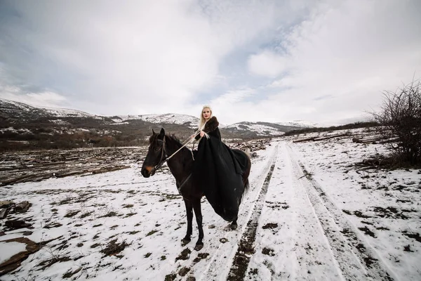 Beautiful blonde Viking in a black cape on horseback