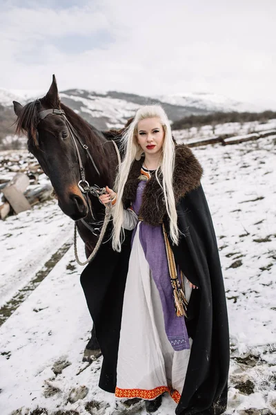 Schöne blonde Wikinger in einem schwarzen Umhang zu Pferd — Stockfoto