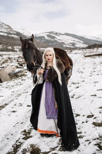 Hermosa rubia vikinga en una capa negra a caballo — Foto de Stock