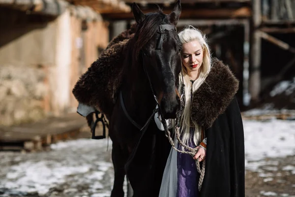 Portrait of a young blond woman in a black cloak with a horse.