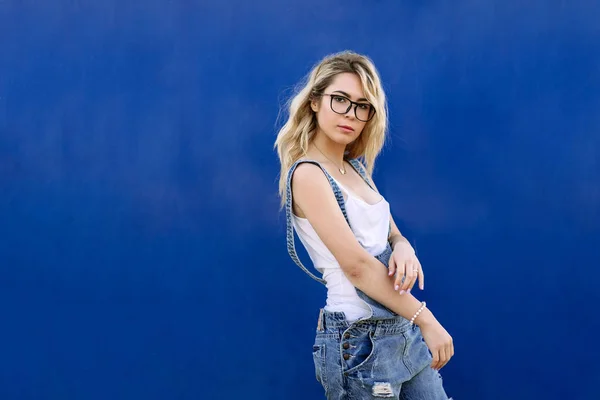 Jovem menina loira elegante em macacões jeans feitos de ganga e em um fundo azul — Fotografia de Stock