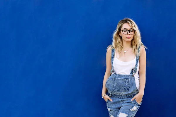 Молодая стильная блондинка в джинсовом комбинезоне из джинса и на синем фоне — стоковое фото