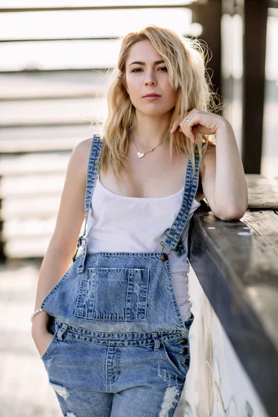 Mladé stylové blondýnka v džínové overaly venku s přirozeným denním světlem, slunečný den — Stock fotografie