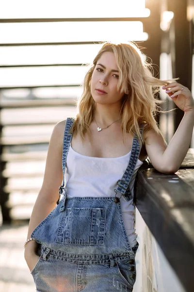 Mladé stylové blondýnka v džínové overaly venku s přirozeným denním světlem, slunečný den — Stock fotografie