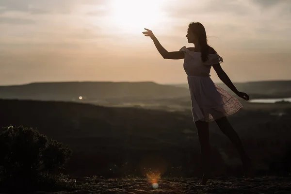 Menina bonita permanece no topo de uma montanha e olha para o horizonte com um belo fundo. Uma foto colorida de um pôr-do-sol natural, um milagre, incrível, uma dança, cabelo ao vento — Fotografia de Stock