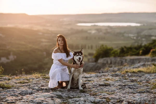 美しい少女は日没で山の犬 (黒と白青い目のハスキー) と遊ぶ — ストック写真