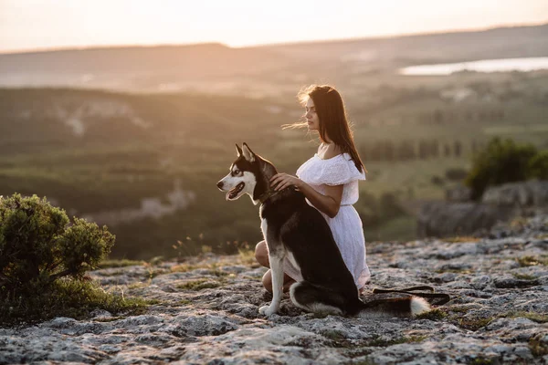 美しい少女は日没で山の犬 (黒と白青い目のハスキー) と遊ぶ — ストック写真