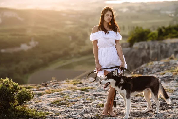 美しい少女は日没で山の犬 (黒と白青い目のハスキー) と遊ぶ — ストック写真