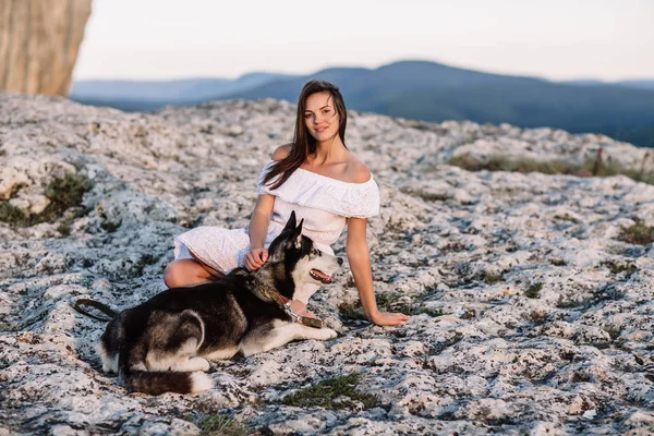 美しい少女は日没で山の犬 (黒と白青い目のハスキー) と遊ぶ — ストック写真