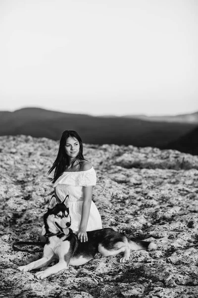 Menina bonita brinca com um cão (preto e branco husky com olhos azuis) nas montanhas ao pôr do sol — Fotografia de Stock