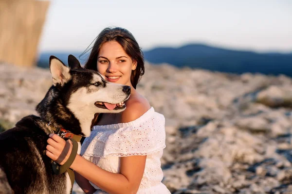 美しい少女は日没で山の犬 (黒と白青い目のハスキー) と遊ぶ — ストック写真