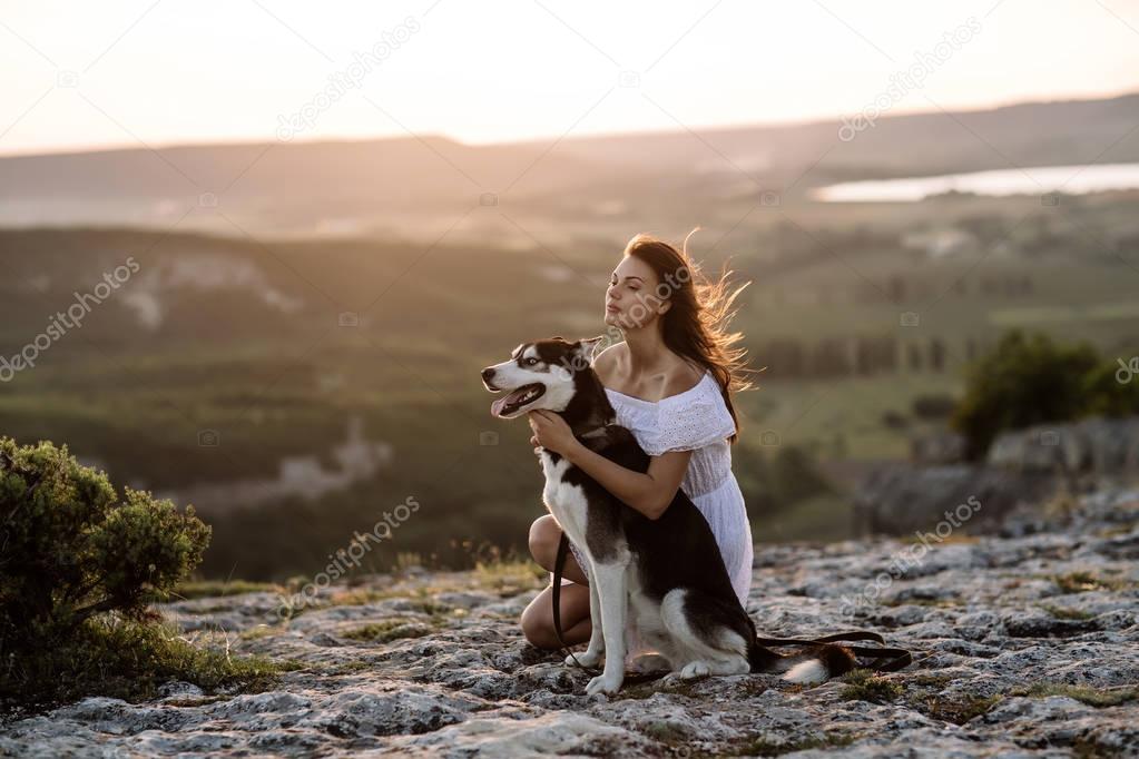 Beautiful girl plays with a dog (black and white husky with blue eyes) in the mountains at sunset