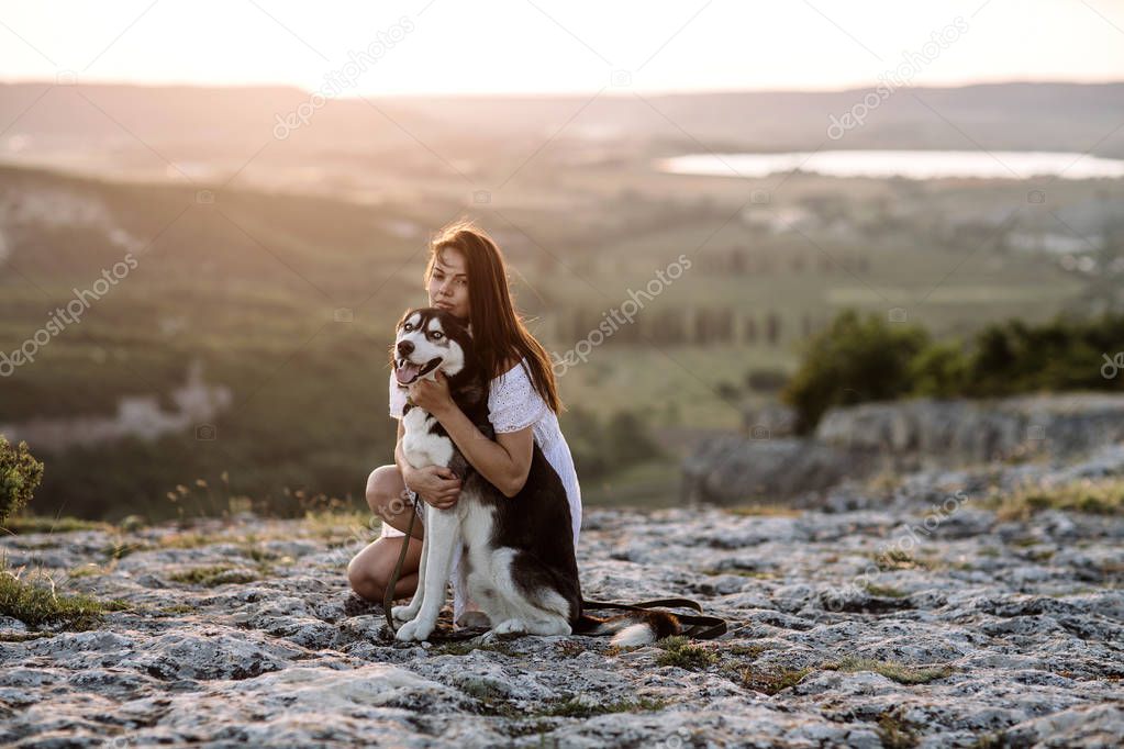 Beautiful girl plays with a dog (black and white husky with blue eyes) in the mountains at sunset