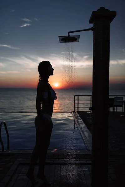 Silueta de una hermosa chica con una figura perfecta en la ducha al atardecer en el mar y la piscina —  Fotos de Stock