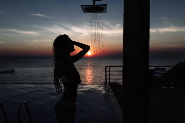 Silhouette di una bella ragazza con una figura perfetta sotto la doccia al tramonto nel mare e in piscina — Foto Stock