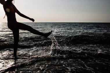 Yaz aylarında güneşli bir günde bacaklarını plajda su ile çalışan kız arkadan aydınlatmalı vurdu. su sıçraması