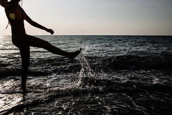 Yaz aylarında güneşli bir günde bacaklarını plajda su ile çalışan kız arkadan aydınlatmalı vurdu. su sıçraması