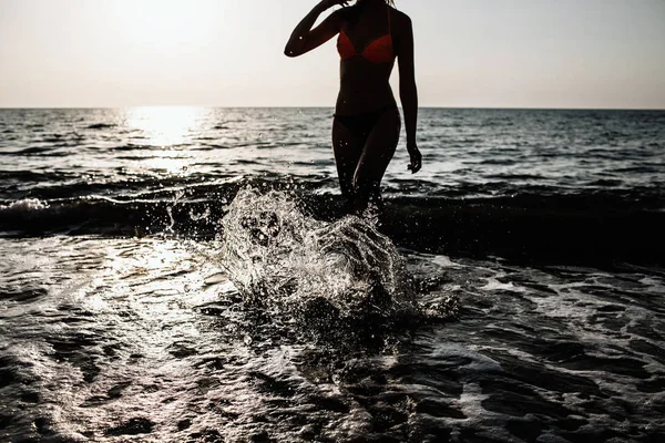 Foto retroiluminada de la chica corriendo con las piernas en el agua en la playa en un día soleado en el verano. salpicaduras de agua —  Fotos de Stock