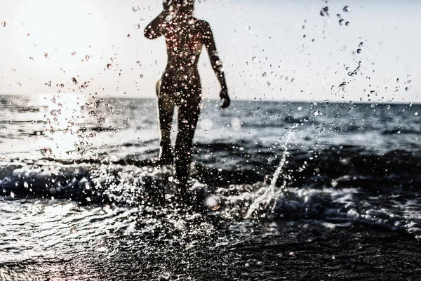 Prise de vue rétroéclairée de la fille courant les jambes dans l'eau à la plage par une journée ensoleillée en été. éclaboussures d'eau — Photo