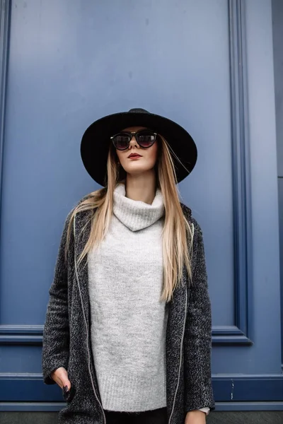 Primo piano ritratto di una giovane bella donna alla moda che indossa occhiali da sole. Un modello in un elegante cappello a tesa larga su sfondo blu. Vestiti armoniosamente simili nei toni del grigio. Stile di Uleach di — Foto Stock