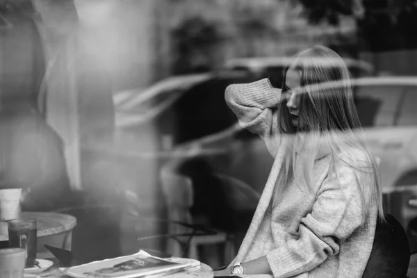 La ragazza a un tavolo in un caffè beve il caffè, il tè, guarda fuori dalla finestra, è colpita attraverso vetro, moda — Foto Stock