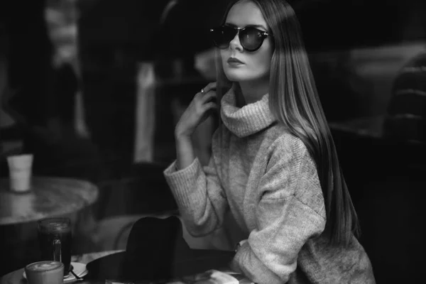 La muchacha a la mesa en la cafetería bebe el café, el té, mira por la ventana, se dispara a través del cristal, la moda — Foto de Stock