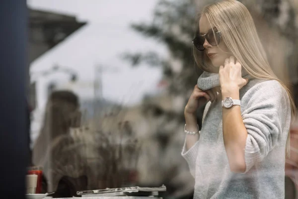 La fille à une table dans un café boit du café, du thé, regarde par la fenêtre, est filmée à travers le verre, la mode — Photo