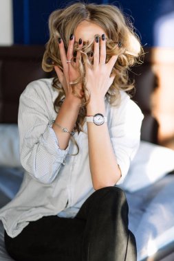 Uzun sarı saçlı bir kız siyah kot pantolon, çizgili bir gömlek ve mavi keten ile yatağa oturmuş Siyah ayakkabılar giymiş. Şık casual kıyafet.