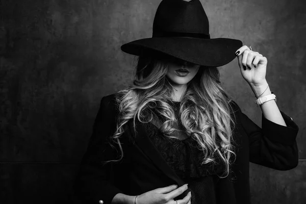 Beautiful young mysterious blond girl in black hat and black jacket on gray background. Eyes are covered with a hat. Manicure - long red nails, nail polish. Fashion, beauty. — Stock Photo, Image