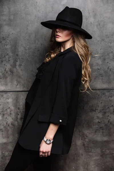 Prachtige mysterieuze blonde meisje in een zwarte hoed en een zwarte jas op grijze achtergrond. Ogen zijn bedekt met een hoed. Manicure - lange rode nagels, nagellak. Mode, schoonheid. — Stockfoto