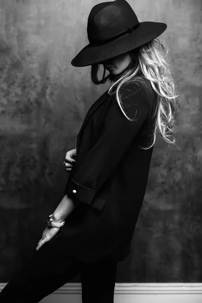 Beautiful young mysterious blond girl in black hat and black jacket on gray background. Eyes are covered with a hat. Manicure - long red nails, nail polish. Fashion, beauty. — Stock Photo, Image