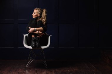 Seksi genç model beyaz bir sandalyede oturuyor ve koyu mavi bir arka plan üzerinde poz koyu giysiler içinde. Yandan dikkat et. Moda, güzellik, stil