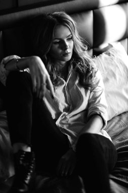 Uzun sarı saçlı bir kız siyah kot pantolon, çizgili bir gömlek ve mavi keten ile yatağa oturmuş Siyah ayakkabılar giymiş. Şık casual kıyafet.