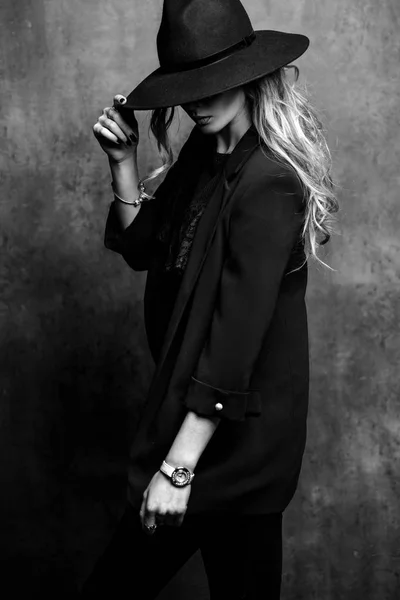 Beautiful young mysterious blond girl in black hat and black jacket on gray background. Eyes are covered with a hat. Manicure - long red nails, nail polish. Fashion, beauty. — Stock Photo, Image