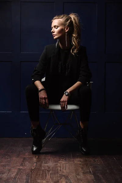 Sexy junges Model in dunkler Kleidung, sitzt auf einem weißen Stuhl und posiert auf dunkelblauem Hintergrund. Uhr zur Hand. Mode, Schönheit, Stil — Stockfoto