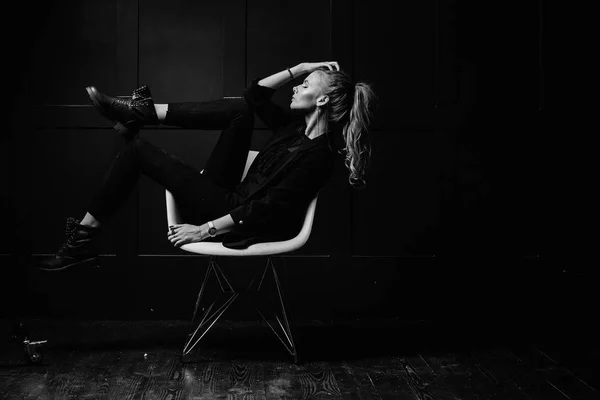 Sexy giovane modella in abiti scuri, seduta su una sedia bianca e in posa su uno sfondo blu scuro. Guarda a portata di mano. Moda, Bellezza, Stile — Foto Stock