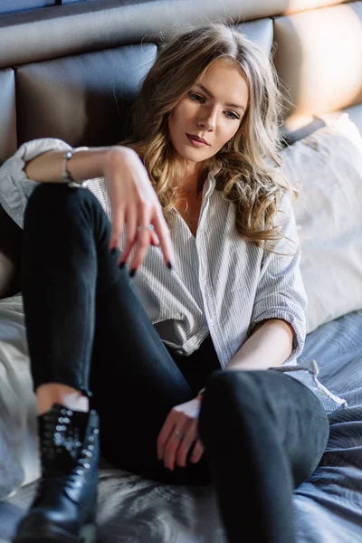 Ein Mädchen mit langen blonden Haaren, bekleidet mit schwarzen Jeans, gestreiftem Hemd und schwarzen Schuhen, die auf dem Bett mit blauer Bettwäsche sitzen. modisch lässiges Outfit. — Stockfoto