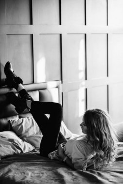 Uzun sarı saçlı bir kız siyah kot pantolon, çizgili bir gömlek ve mavi keten ile yatağa oturmuş Siyah ayakkabılar giymiş. Şık casual kıyafet.