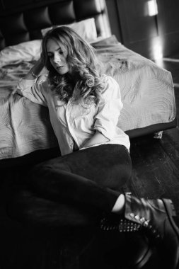 Uzun sarı saçlı bir kız siyah kot pantolon, çizgili bir gömlek ve mavi keten ile yatağa oturmuş Siyah ayakkabılar giymiş. Şık casual kıyafet.