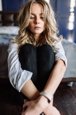 Uzun sarı saçlı bir kız siyah kot pantolon, çizgili bir gömlek ve mavi keten ile yatağa oturmuş Siyah ayakkabılar giymiş. Şık casual kıyafet.