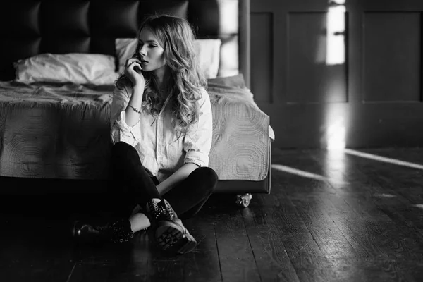Ένα κορίτσι με μακριά ξανθά μαλλιά, ντυμένοι με ένα ριγέ πουκάμισο, μαύρο τζιν και μαύρα παπούτσια κάθεται στο κρεβάτι με μπλε σεντόνια. Μοντέρνα casual ντύσιμο. — Φωτογραφία Αρχείου