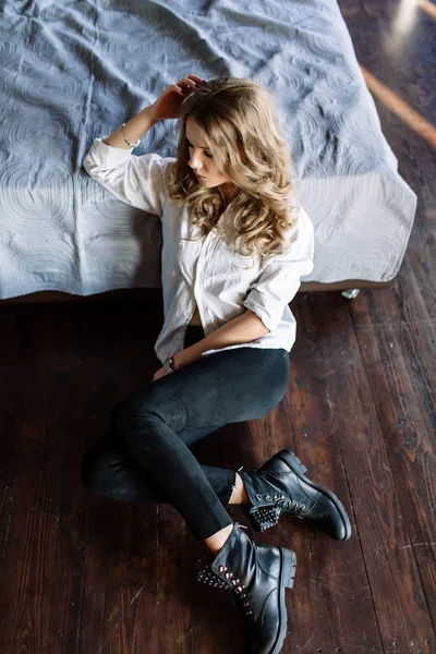 Ein Mädchen mit langen blonden Haaren, bekleidet mit schwarzen Jeans, gestreiftem Hemd und schwarzen Schuhen, die auf dem Bett mit blauer Bettwäsche sitzen. modisch lässiges Outfit. — Stockfoto