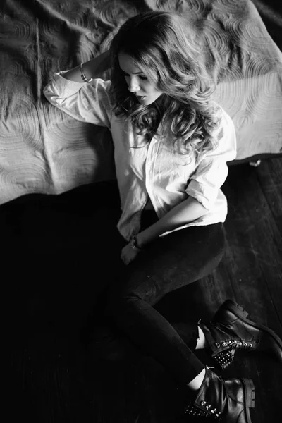 A girl with long blond hair dressed in black jeans, a striped shirt and black shoes sitting on the bed with blue linens. Fashionable casual outfit. — Stock Photo, Image