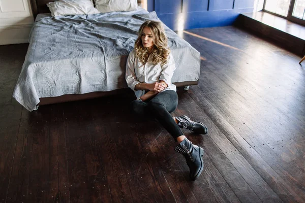 Dziewczyna z długimi blond włosami, ubrany w czarne dżinsy, pasiastą koszulę i czarne buty siedzi na łóżku z niebieski pościel. Modny strój casual. — Zdjęcie stockowe