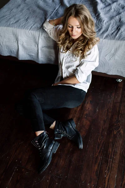 Ein Mädchen mit langen blonden Haaren, bekleidet mit schwarzen Jeans, gestreiftem Hemd und schwarzen Schuhen, die auf dem Bett mit blauer Bettwäsche sitzen. modisch lässiges Outfit. — Stockfoto