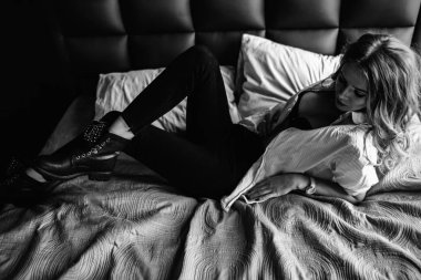 Uzun sarı saçlı bir kız siyah kot pantolon, çizgili bir gömlek ve mavi keten ile yatağa oturmuş Siyah ayakkabılar giymiş. Şık casual kıyafet.