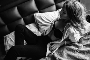 Uzun sarı saçlı bir kız siyah kot pantolon, çizgili bir gömlek ve mavi keten ile yatağa oturmuş Siyah ayakkabılar giymiş. Şık casual kıyafet.