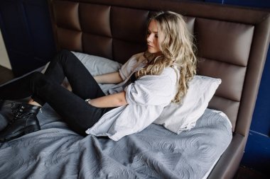 Uzun sarı saçlı bir kız siyah kot pantolon, çizgili bir gömlek ve mavi keten ile yatağa oturmuş Siyah ayakkabılar giymiş. Şık casual kıyafet.