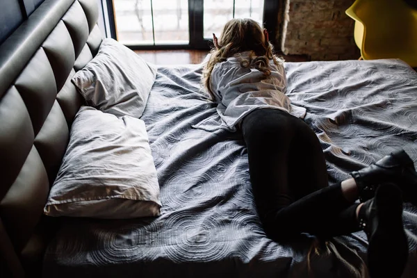 Une fille aux longs cheveux blonds vêtus de jeans noirs, une chemise rayée et des chaussures noires assises sur le lit avec des draps bleus. Tenue casual à la mode . — Photo