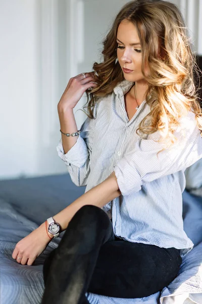 Uma menina com longos cabelos loiros vestida com jeans pretos, uma camisa listrada e sapatos pretos sentados na cama com lençóis azuis. Roupa casual na moda . — Fotografia de Stock